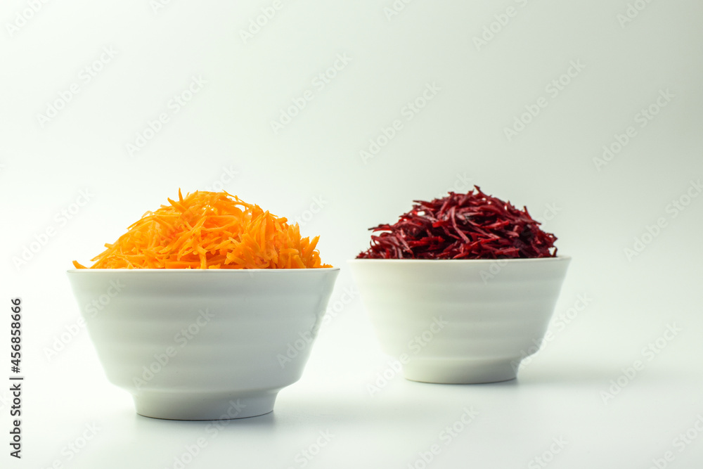 Fresh grated carrot and beet in white bowls, on a white background. Selective focus. Side view. Fresh grated carrot and beet in white bowls, on a white background. Selective focus. Side view.