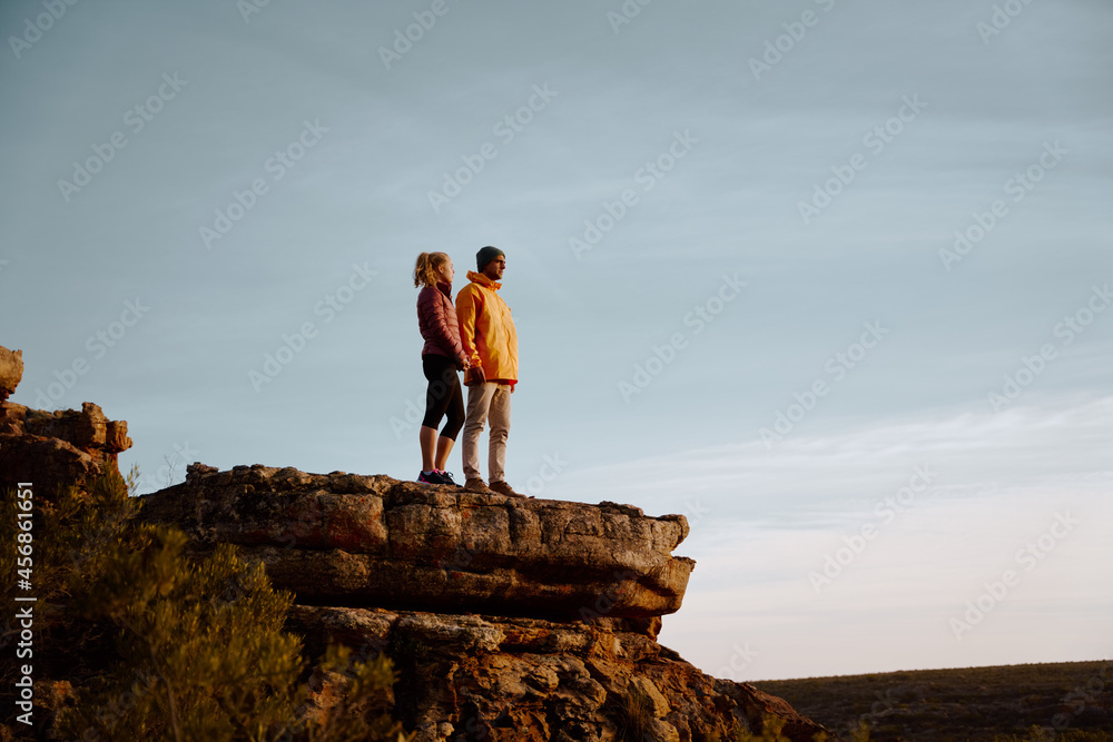 Young couple holding hands looking away standing at peak of cliff after successful hike