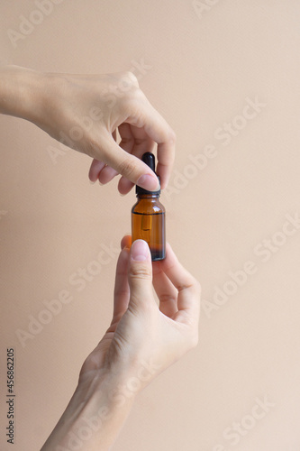 Women's hands hold mock-up of glass dropper bottle and unscrew lid, on beige background. Vertical image, close-up. Concept of body care, spa treatments, natural cosmetics