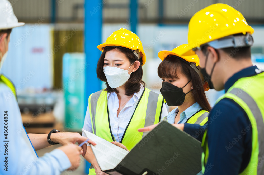 The factory employee consists of engineers, foreman, technicians, and related department staff. Wear a mask, hard hat, and vest. meeting before starting work inside the warehouse. Teamwork concept. The factory employee consists of engineers, foreman, technicians, and related department staff. Wear a mask, hard hat, and vest. meeting before starting work inside the warehouse. Teamwork concept.