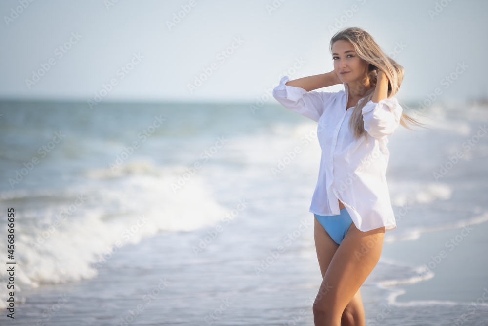 A girl with long hair in a bluish swimsuit and a shirt goes through her hair on the seashore