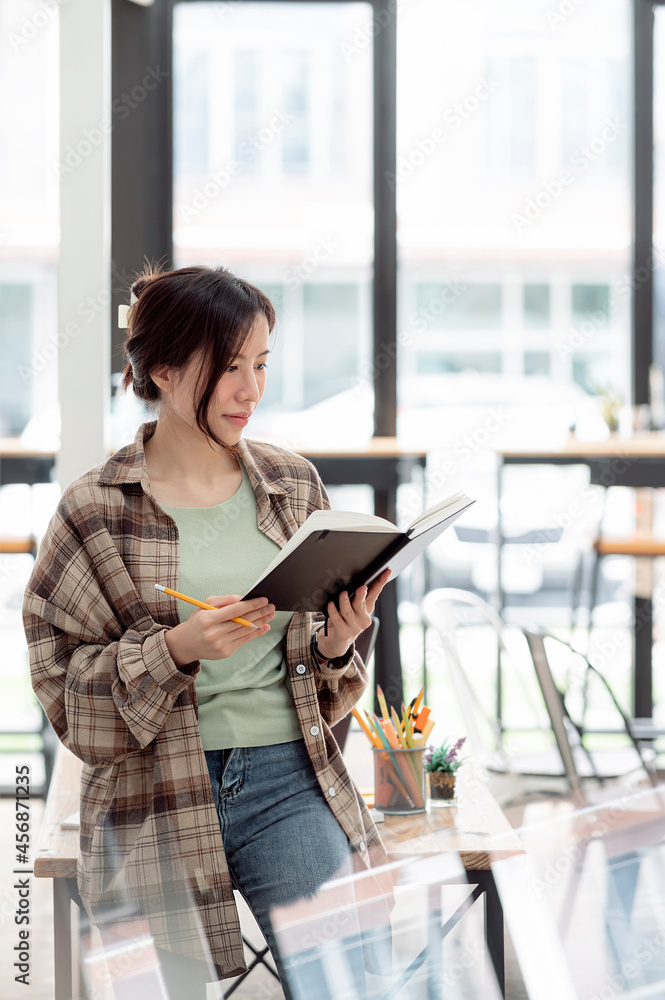 Fototapeta premium Portrait of young asian woman looking at notebook in her hand while standing at office room, vertical view.