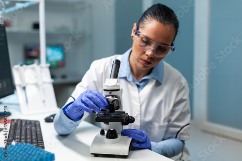 Biologist researcher working at vaccine development against virus putting blood sample under medical microscope during microbiology experiment. Chemist doctor sitting at table in hospital laboratory