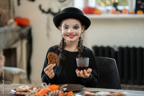 Happy girl in halloween attire having cookie and drink while sitting by festive table