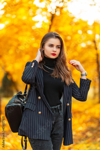 Elegant beautiful young woman model with red lips in a fashionable black blazer and sweater with a leather backpack walks in the park with yellow autumn foliage. Business female style outdoors