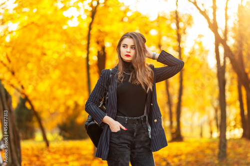 Beautiful young woman with red lips in a fashion elegant suit with a blazer, sweater and backpack walks in the park with colorful yellow-orange foliage at sunset