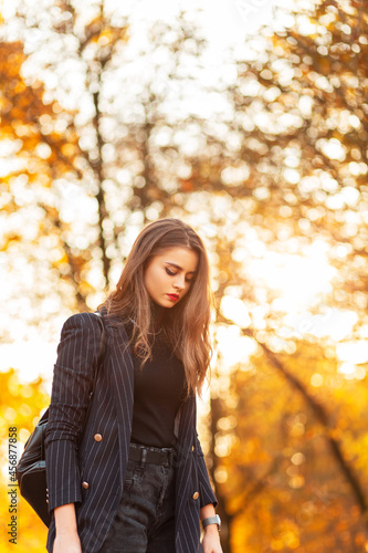beautiful girl in a trendy business suit with a stylish blazer and a sweater with a fashion backpack walks in the park with golden autumn foliage at sunset