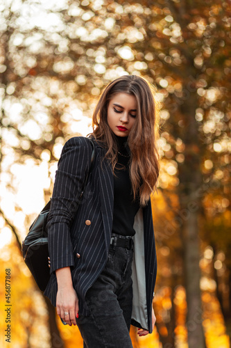Elegant beautiful business woman with red lips in a fashion black suit with blazer and a leather backpack walks in a park with yellow autumn foliage