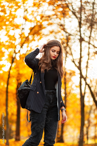 Stylish beautiful young woman model in a black business suit with a fashionable blazer, sweater and backpack walks in an autumn park with yellow autumn foliage at sunset