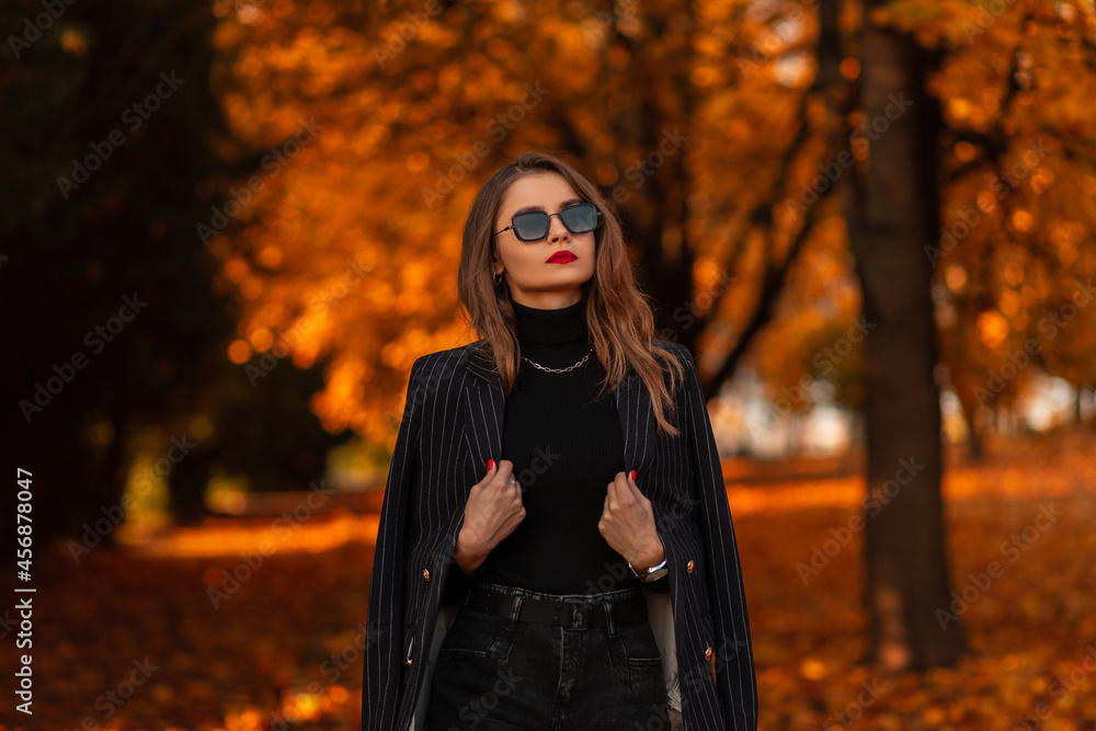 Portrait of a beautiful fashionable girl with sunglasses in a fashion black suit with a blazer and a sweater walks in the park with colorful golden foliage Portrait of a beautiful fashionable girl with sunglasses in a fashion black suit with a blazer and a sweater walks in the park with colorful golden foliage