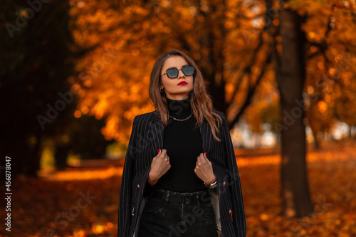 Portrait of a beautiful fashionable girl with sunglasses in a fashion black suit with a blazer and a sweater walks in the park with colorful golden foliage