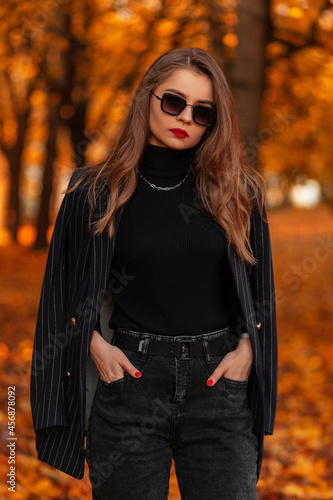 Fashionable woman with red lips in fashion black elegant suit with blazer and sweater with sunglasses in park with yellow autumn foliage