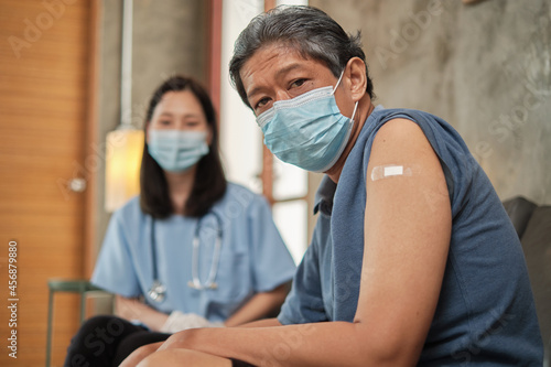 An elderly Asian male patient has a plaster bandage on his arm after vaccinating against COVID-19. This is a medical service from a specialist female doctor at a senior retirement home.