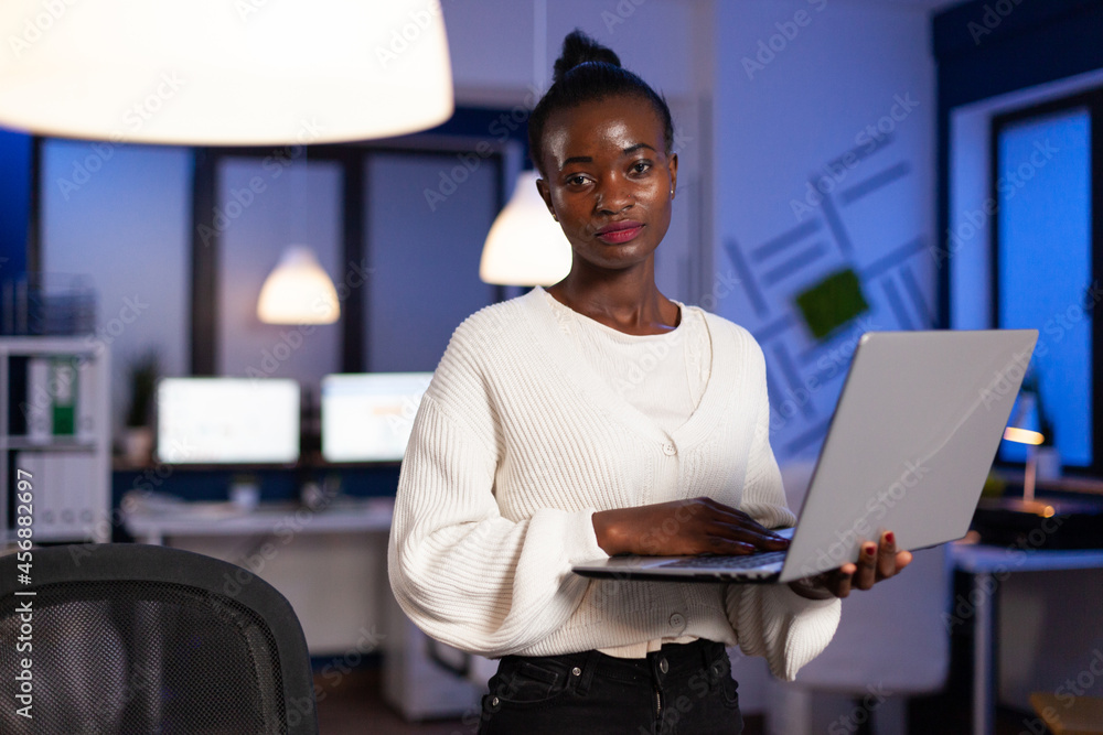 Portrait of african american executive manager holding laptop computer while browsing online business information late at night in company office. Black businesswoman hardworking at financial strategy