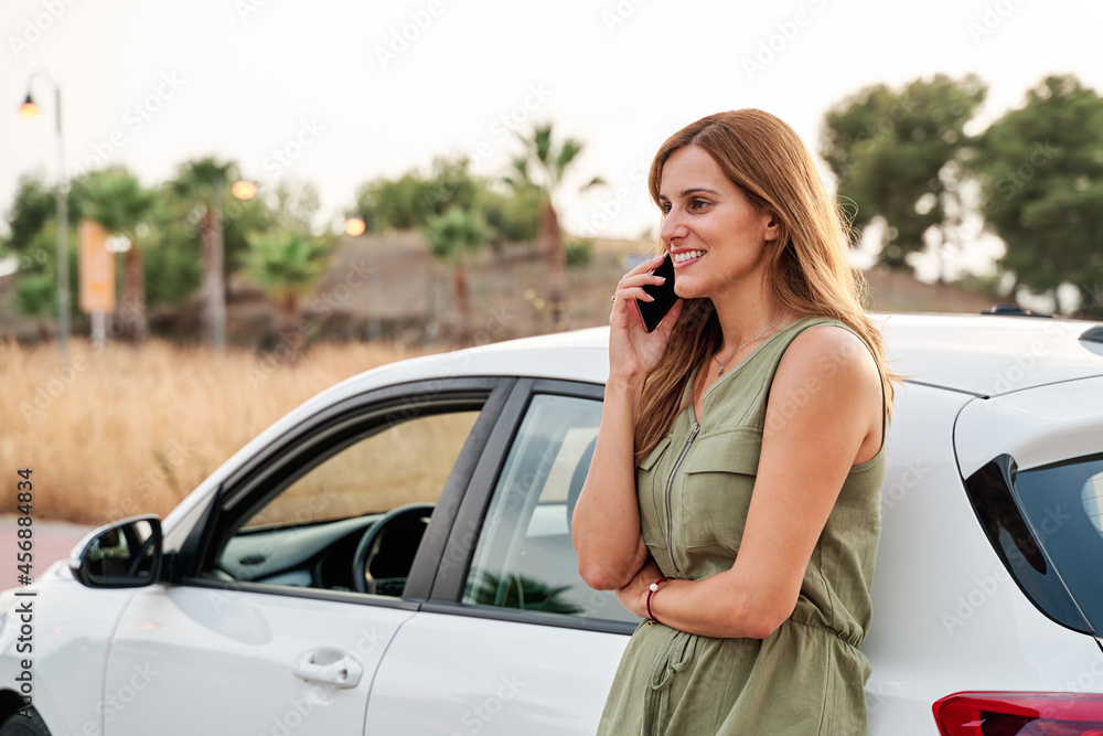Young pretty woman standing leaning against her car while talking on her smart phone.