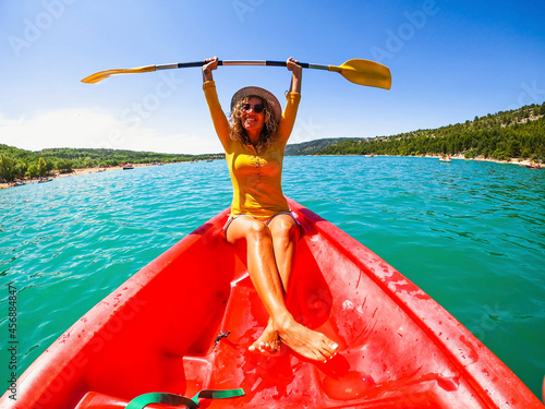Happy caucasian woman holding oar sitting on red canoe in lake during sunny day. Beautiful female tourist holding oar with both arms raised having fun on a canoe ride during summer