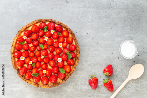 French strawberry tart with wild strawberry leaves and flowers on top of a cooling rack with sugar and spoon on a grey background. Flat Lay, top view.