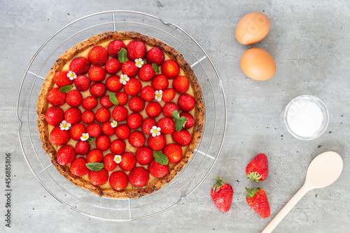 French strawberry tart with wild strawberry leaves and flowers on top of a cooling rack with eggs, sugar and spoon on a grey background. Flat Lay, top view.