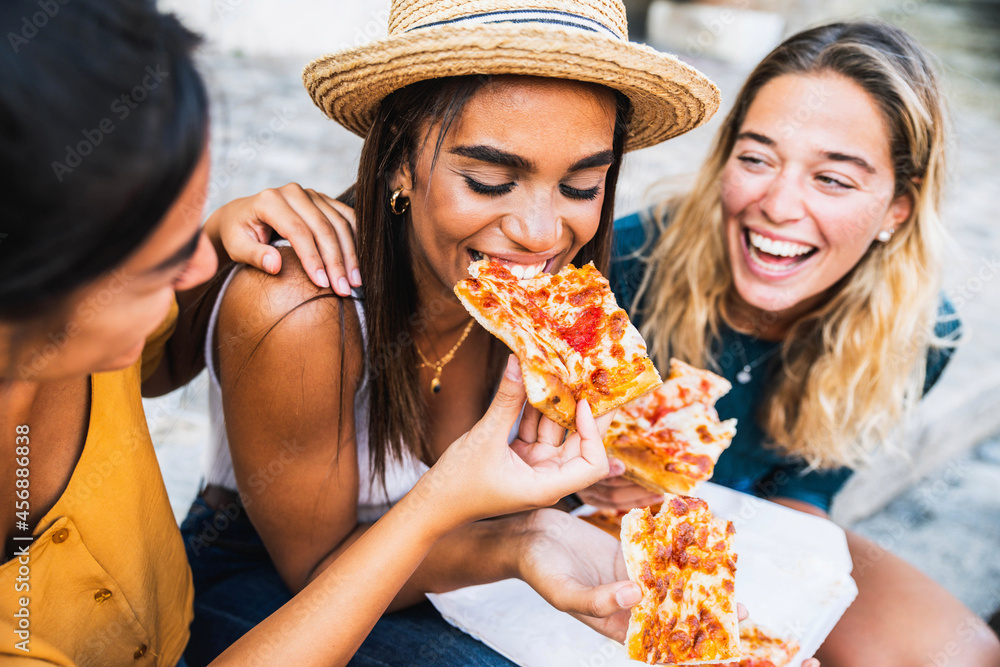 © Davide Angelini - Three young female friends sitting outdoor and eating pizza - Happy women having fun enjoying a day out on city street - Happy lifestyle concept