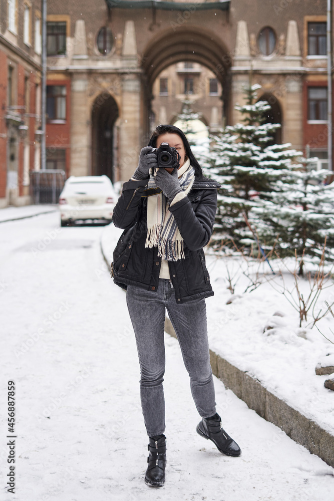 a young woman in winter holds a camera near her face and takes pictures in a European city. there is snow around