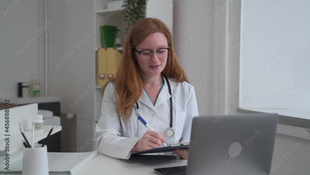 Doctor ehealth online service. Spbas Attentive woman practitioner with glasses makes notes examining patient at videochat via laptop in clinic office Doctor ehealth online service. Spbas Attentive woman practitioner with glasses makes notes examining patient at videochat via laptop in clinic office
