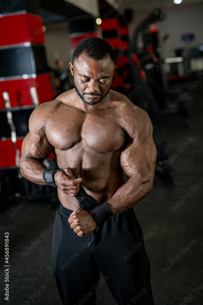 Vertical view of the young multiracial man with naked torso putting special bandages at his hands on modern gym background. Strength and motivation concept