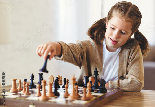 Cute little girl playing chess while spending leisure time at home during during day time, child sitting at table in living room and moving figure on chessboard with concentrated face expression