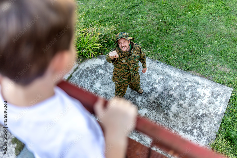 Fototapeta premium Happy soldier reunited with his son after coming back from war. Excited boy standing on terrace with open arms for hug, looking at military dad in camouflage. Veteran of war or returning home concept.
