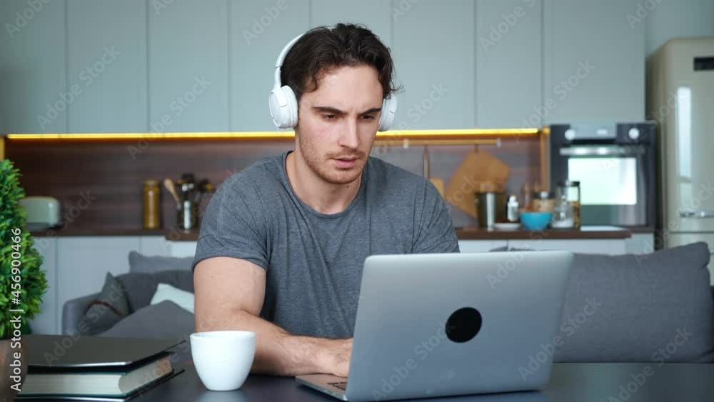 Handsome young man, attractive guy , freelance employee, businessman wearing wireless headphones, sitting at the table with cup of coffee and folder of documents, working on laptop remotely from home Handsome young man, attractive guy , freelance employee, businessman wearing wireless headphones, sitting at the table with cup of coffee and folder of documents, working on laptop remotely from home