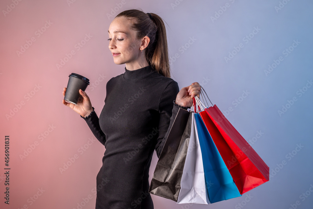 Modern young woman in a black dress is holding cup of coffee and colorful paper bags with purchases from a shop in her hands on a blue pink background. Concept of shopping, sale and Black Friday Modern young woman in a black dress is holding cup of coffee and colorful paper bags with purchases from a shop in her hands on a blue pink background. Concept of shopping, sale and Black Friday