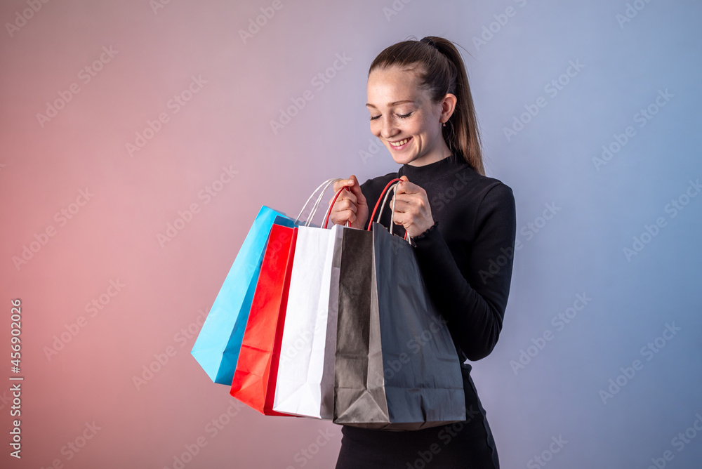Young pretty woman in a black dress is holding colorful paper bags with purchases from the shop in her hands and looking into it with a smile. Concept of shopping, sale and Black Friday. Copy space