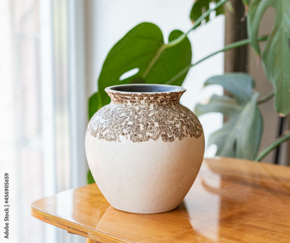 Mid-century modern pottery, beige vase on a wooden table with philodondendron in the background Mid-century modern pottery, beige vase on a wooden table with philodondendron in the background