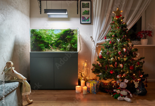 Glowing Christmas tree with aquascape aquarium. Toy rabbit looks aquascape, burning candles and gifts. Rabbit below tree hugs a little elf. photo