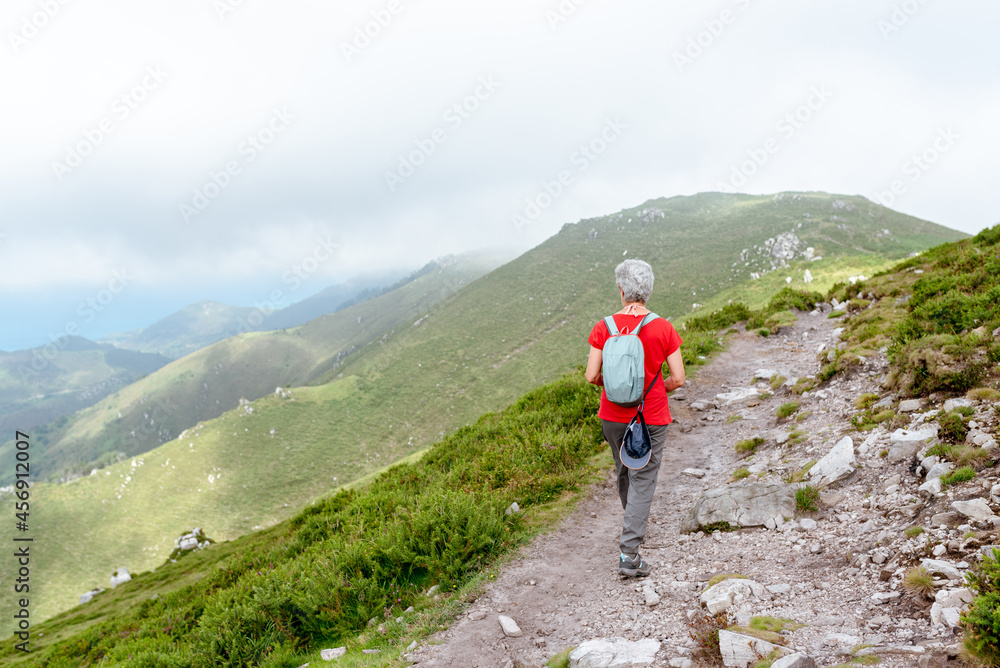 senior woman hiker with backpack and gray hair walking on a mountain path on a cloudy day