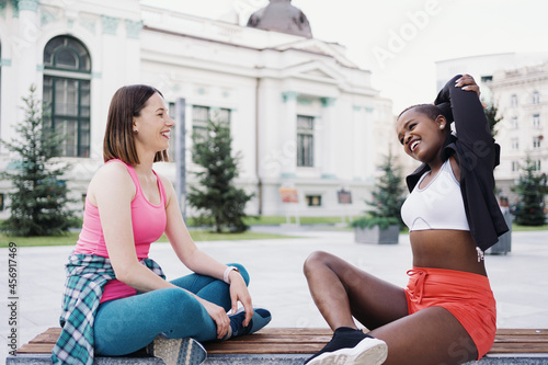 Cheerful smiling friends in sportswear sitting on bench in the city dicussing in park. Multiethnic women having a fitness workout break. photo