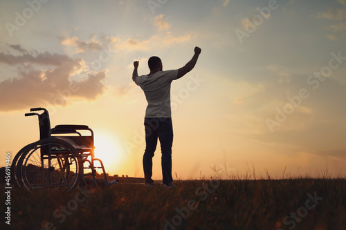 Fotografie Man raising hands up to sky near wheelchair at sunset, back view
