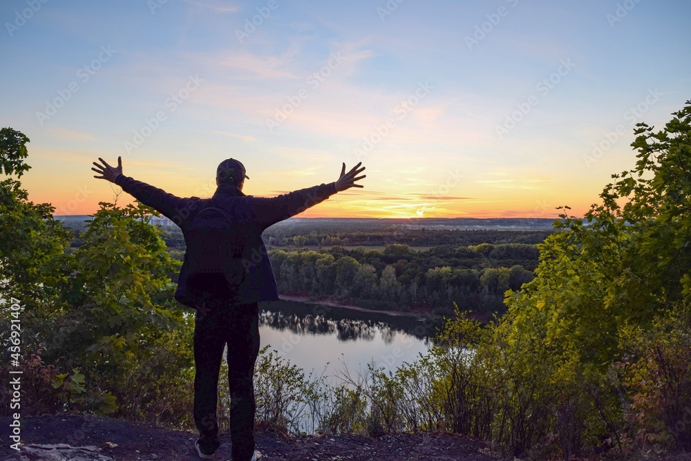 Silhouette of a man with a backpack and arms outstretched against the background of a river and an autumn sunset