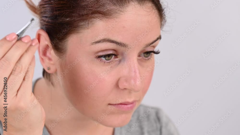 Caucasian woman looks in the mirror and does the eyebrow correction herself with tweezers Caucasian woman looks in the mirror and does the eyebrow correction herself with tweezers
