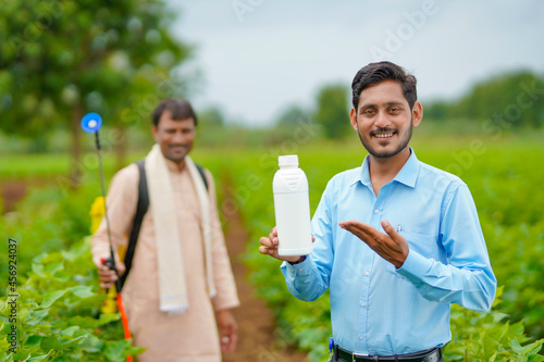 Young indian agronomist giving liquid fertilizer bottle to farmer and saying product information at green agriculture field.