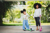 Two playful African sisters with curly hair enjoying and playing while riding scooter on empty lane surrounded by trees during day