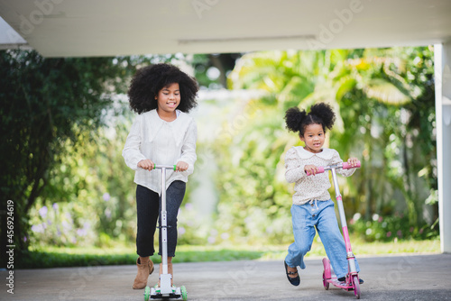 Two playful African sisters with curly hair enjoying and playing while riding scooter on empty lane surrounded by trees during day