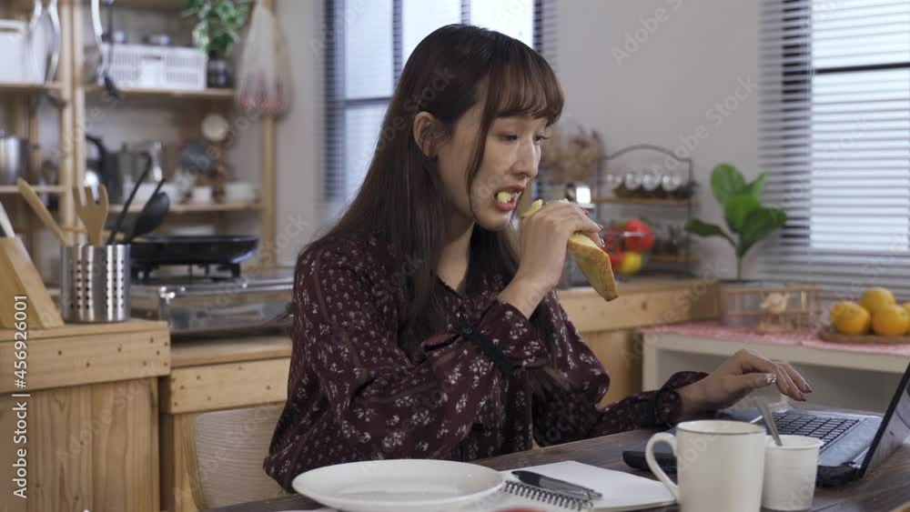 pretty asian manager is eating a toast for breakfast while reading a document on her notebook at the dining table in the morning. pretty asian manager is eating a toast for breakfast while reading a document on her notebook at the dining table in the morning.