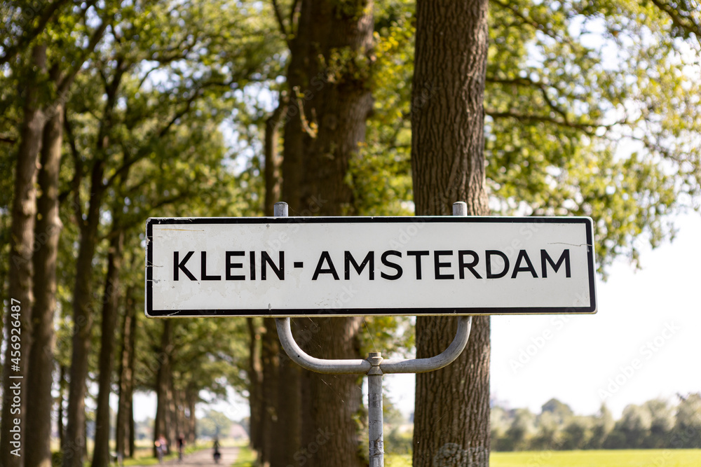 Tree trunks and greenery behind side of the road municipality sign with name of the village Klein Amsterdam you are entering