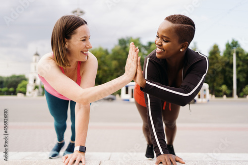 Two smiling diverse young woman in Athletic Workout Clothes are Doing a Plank Exercise working out together in city square. © arthurhidden