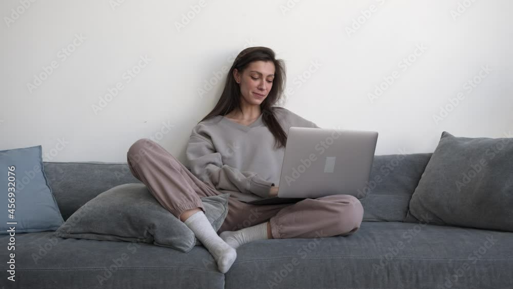 Woman freelancer using device sitting on grey couch, waving hands on a video conference. Gimbal shot of young woman working at home, greet people in online meeting Woman freelancer using device sitting on grey couch, waving hands on a video conference. Gimbal shot of young woman working at home, greet people in online meeting