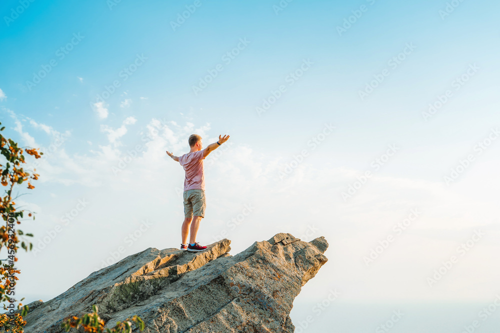 A happy young man with his hands raised stands on a picturesque steep cliff above the sea against the sky. The concept of travel and freedom.