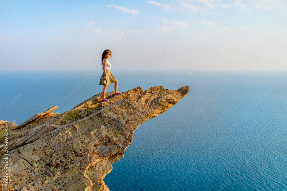 A young woman stands on a picturesque steep cliff above the sea against the sky. The concept of travel and freedom.