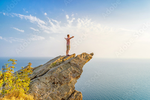 A happy young man with his hands raised stands on a picturesque steep cliff above the sea against the sky. The concept of travel and freedom.