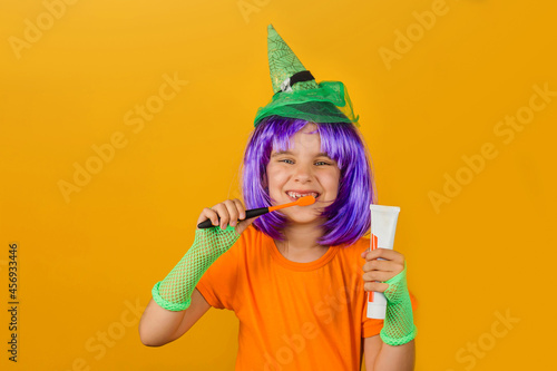 One little girl childin a carnival costume witch with toothpaste and an orange brush for Halloween is isolated on a yellow background. Medicine, dental hygiene, holidays concept.