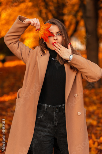 Fresh portrait of a beautiful young woman with emotion of surprise in a fashion beige coat with a bright autumn leaf travel in a park with orange foliage. New collection of fall female look clothes